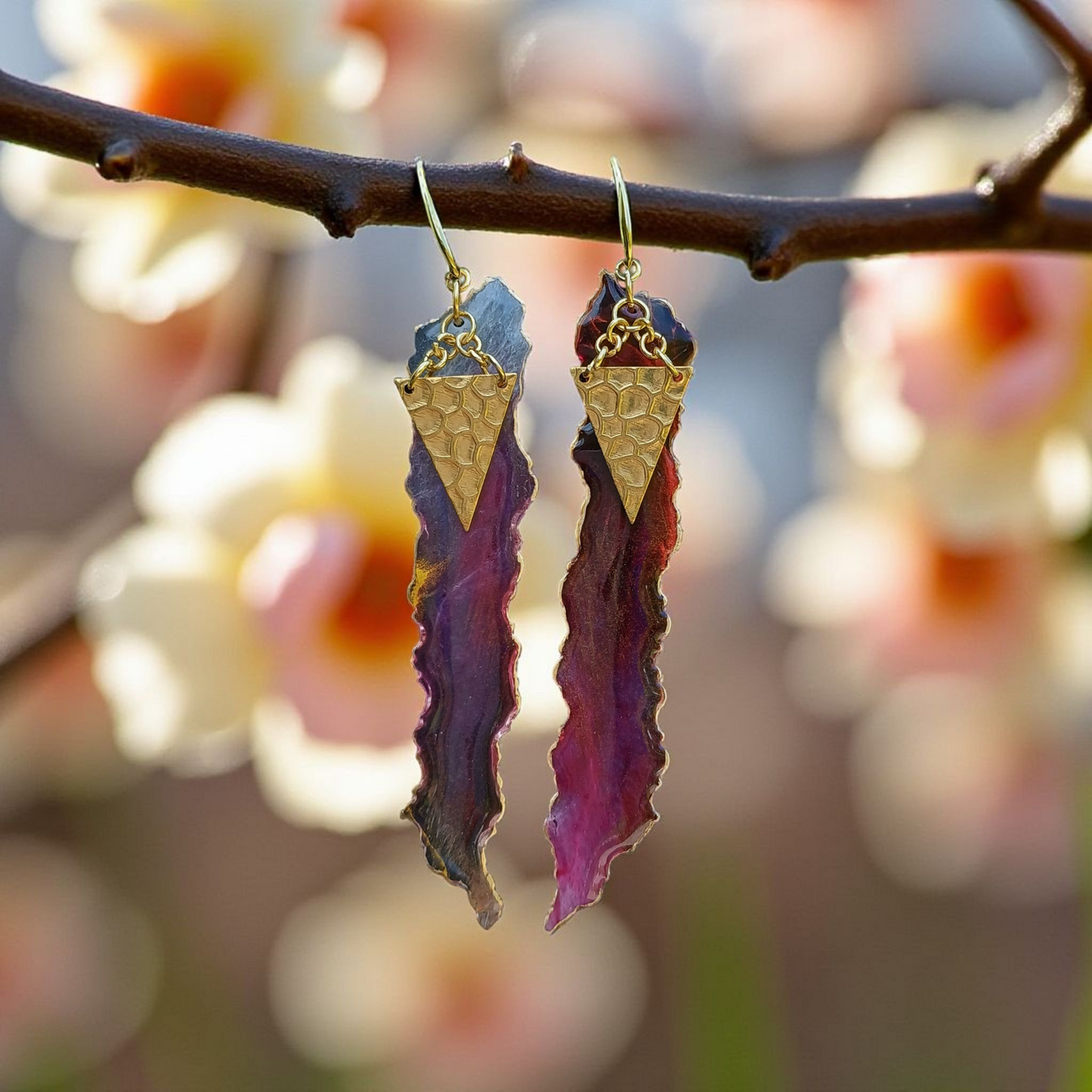 Purple leaf-shaped earrings with gold accents hanging on a branch with blurred floral background