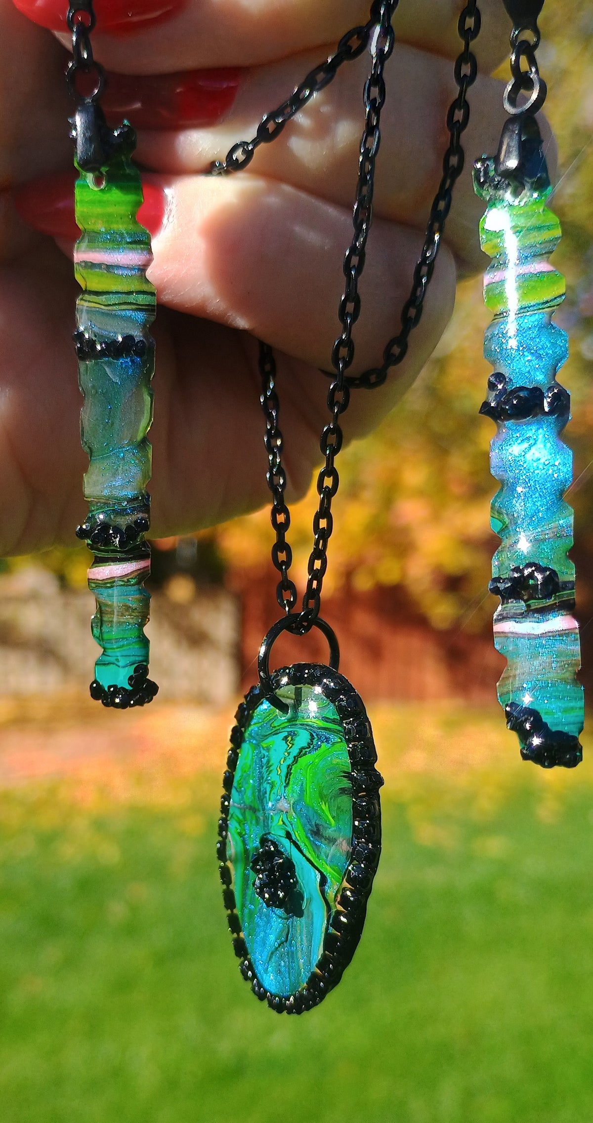 A handmade jewelry set featuring a green and blue marbled pendant necklace and matching drop earrings held by a hand, with a blurred outdoor background a Style By Jess at Gotta Have It Design