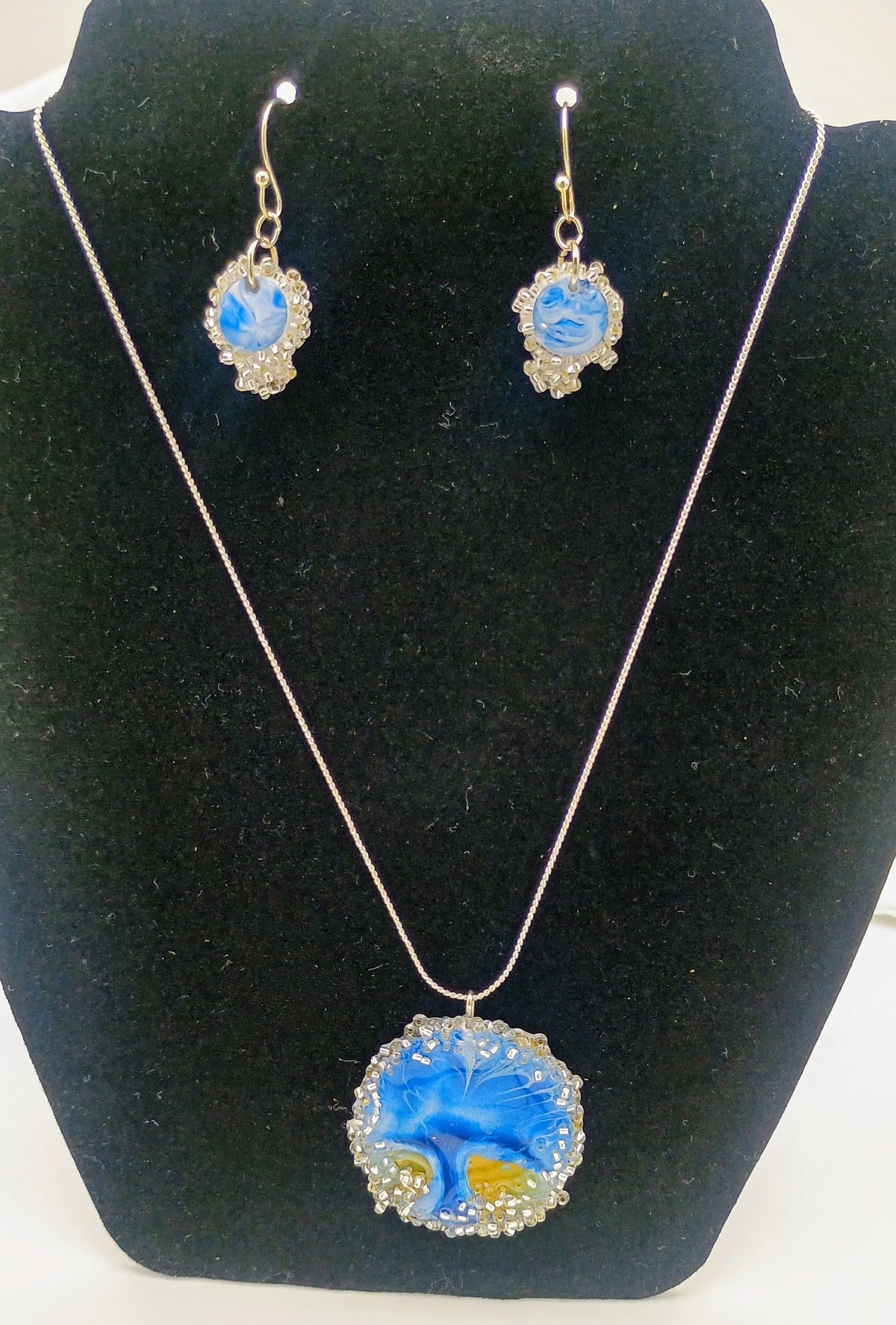 A jewelry set featuring a necklace and matching drop earrings displayed on a black velvet mannequin bust. The set includes a round pendant necklace and matching drop earrings with blue and yellow marbled enamel details surrounded by silver-toned rhinestone borders, suspended from silver-toned chains.