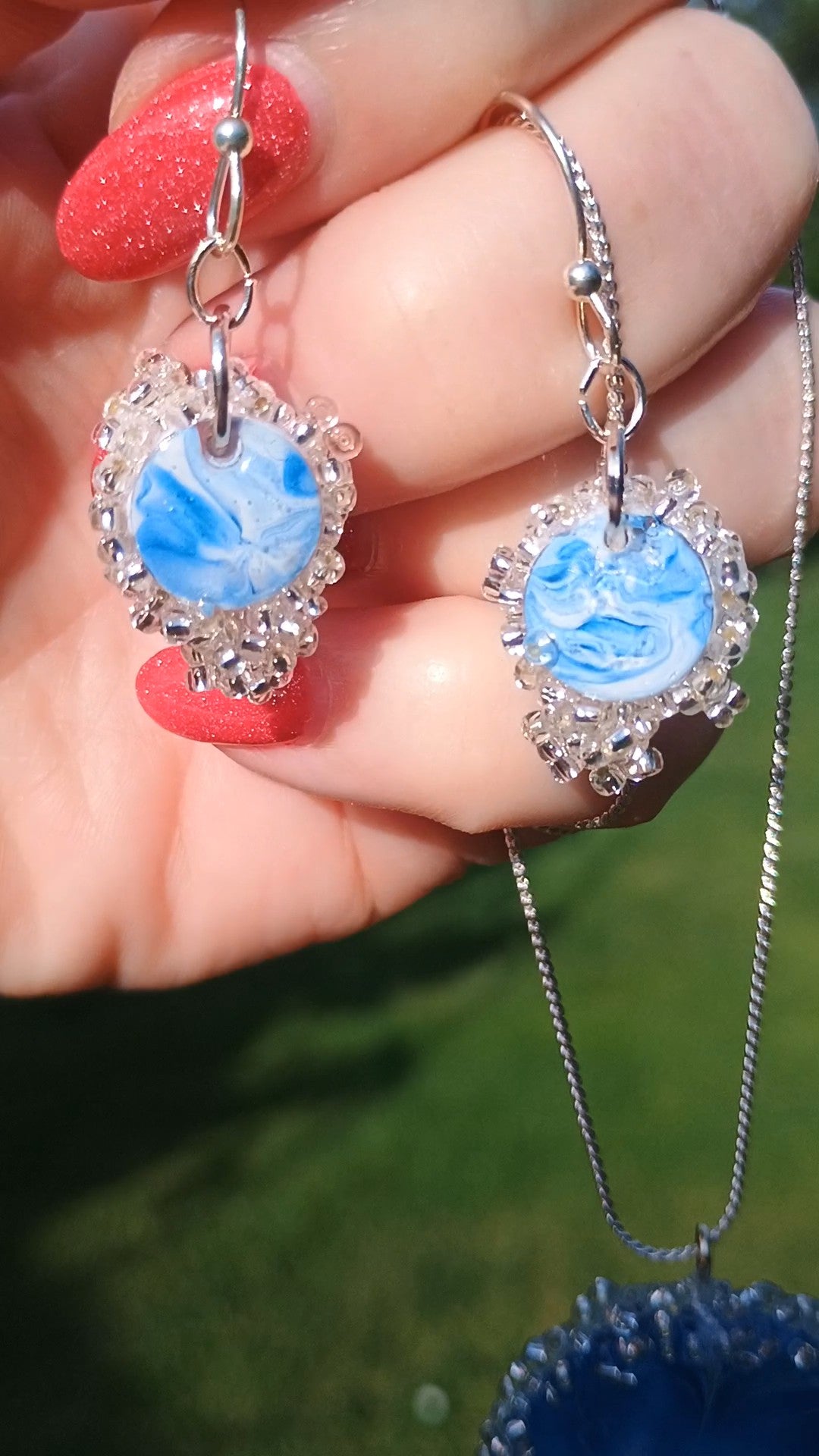 A close-up of a hand with red glittery fingernails holding a pair of dangling earrings and a matching necklace outdoors. The jewelry features round light blue marbled cabochons surrounded by clear rhinestone-like crystals and silver-toned metal settings and chains.
