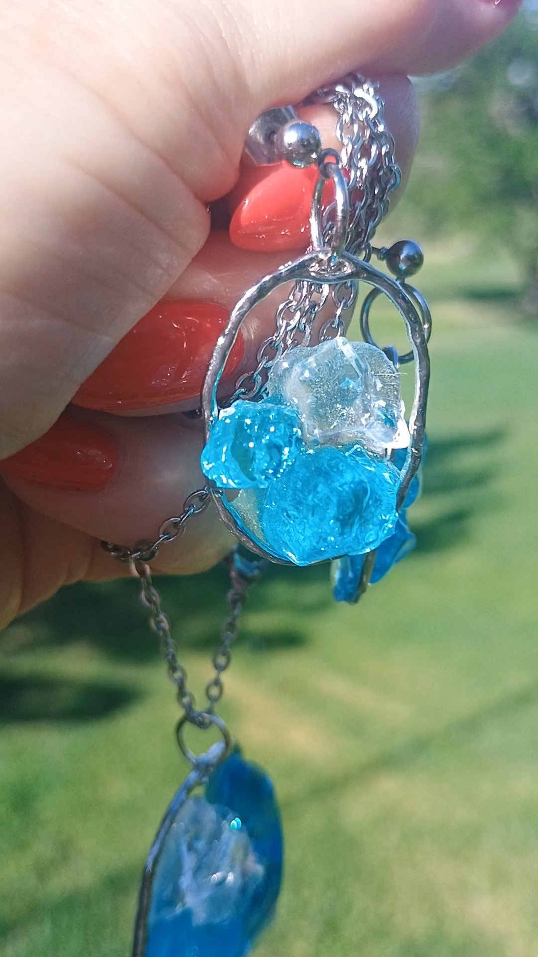 A close-up of a hand holding a silver chain necklace with a blue and clear crystal pendant, set against a blurred green outdoor background. Style By Jess