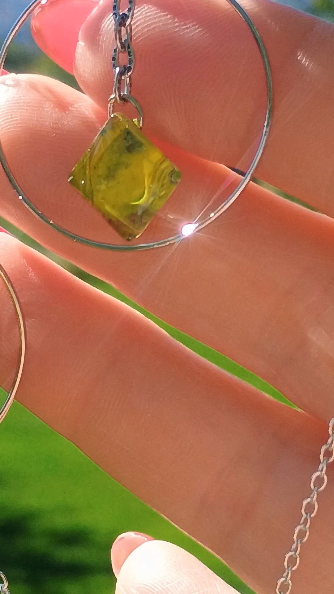 A close-up of a hand holding a silver chain necklace with a small square yellow-green stone pendant, with a large silver hoop also visible.