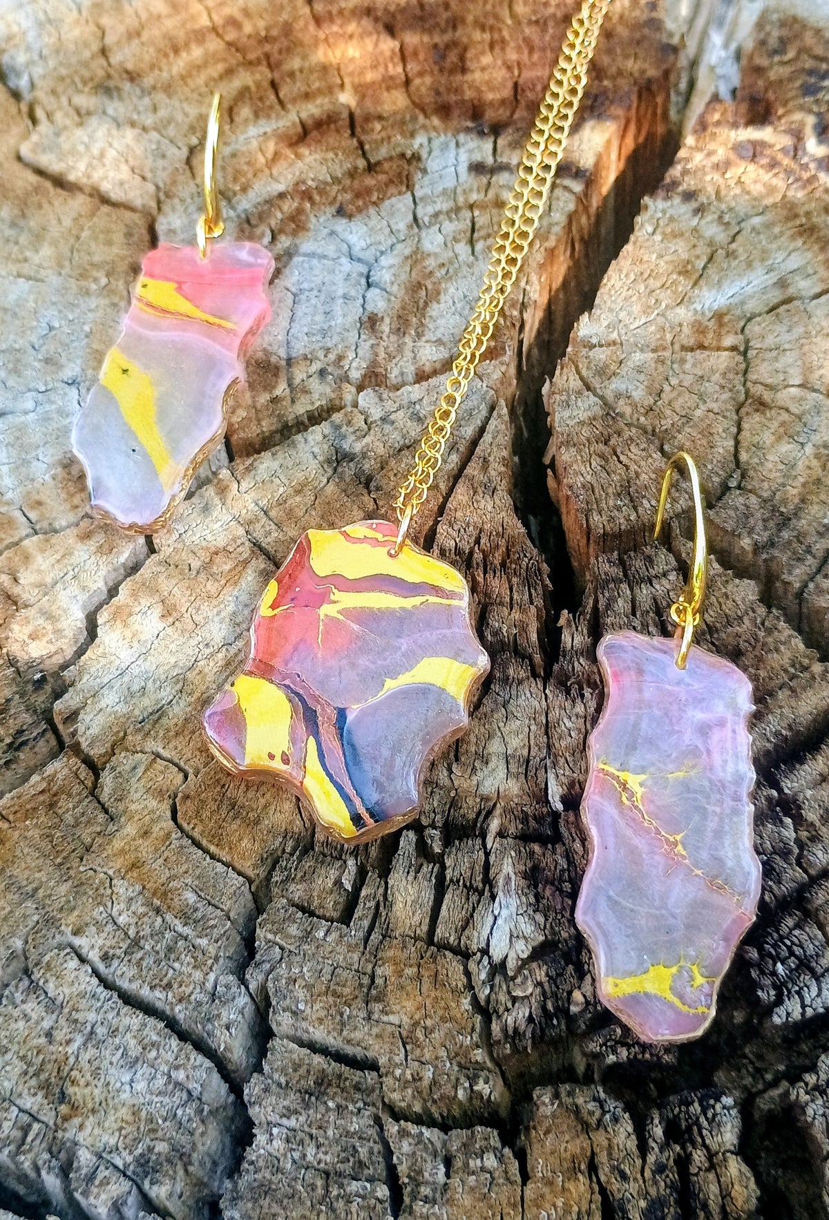 A jewelry set featuring a necklace and two matching drop earrings with irregular marbled stone pendants in pink, purple, yellow, and blue, displayed on a textured wood surface.