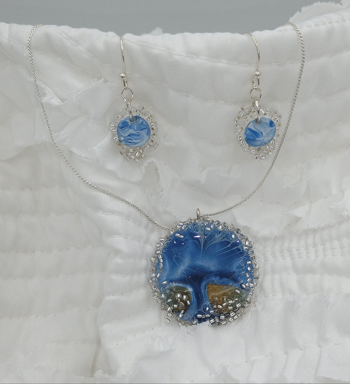 A jewelry set featuring a silver necklace and matching earrings with blue and white marbled stone pendants, displayed on a white ruffled fabric background.