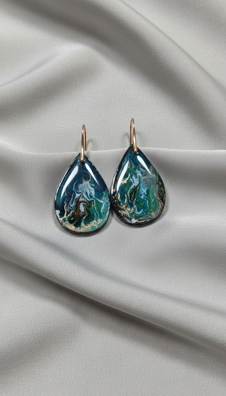 Handmade teal and emerald green resin teardrop dangle earrings with gold glitter accents on 18K gold plated hooks, displayed on white satin fabric — Style By Jess