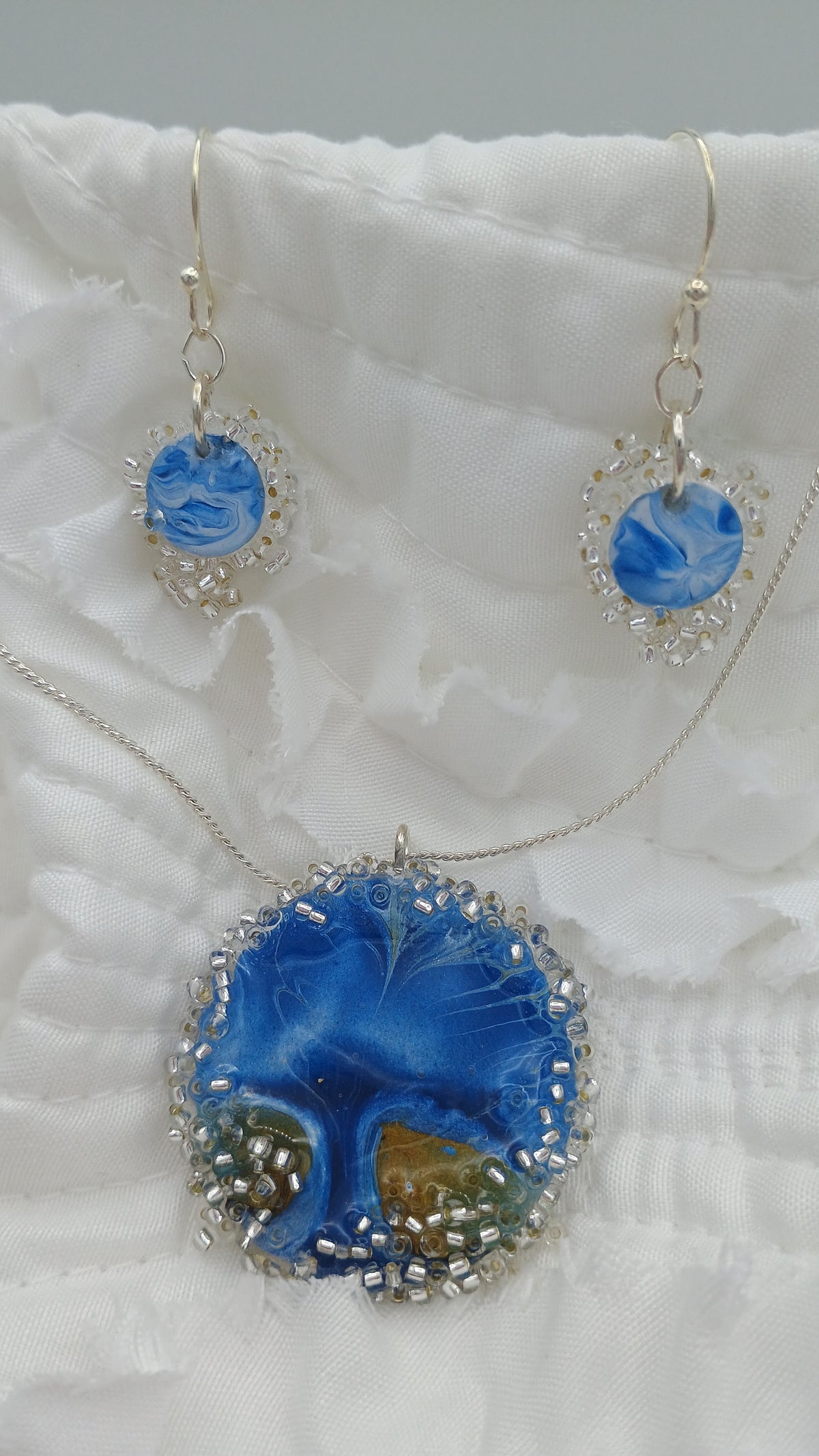 Handmade blue resin dangle earrings with crystal seed bead edges and silver-plated hooks, held in hand outdoors — fluid art jewelry by Style By Jess at Gotta Have It Design