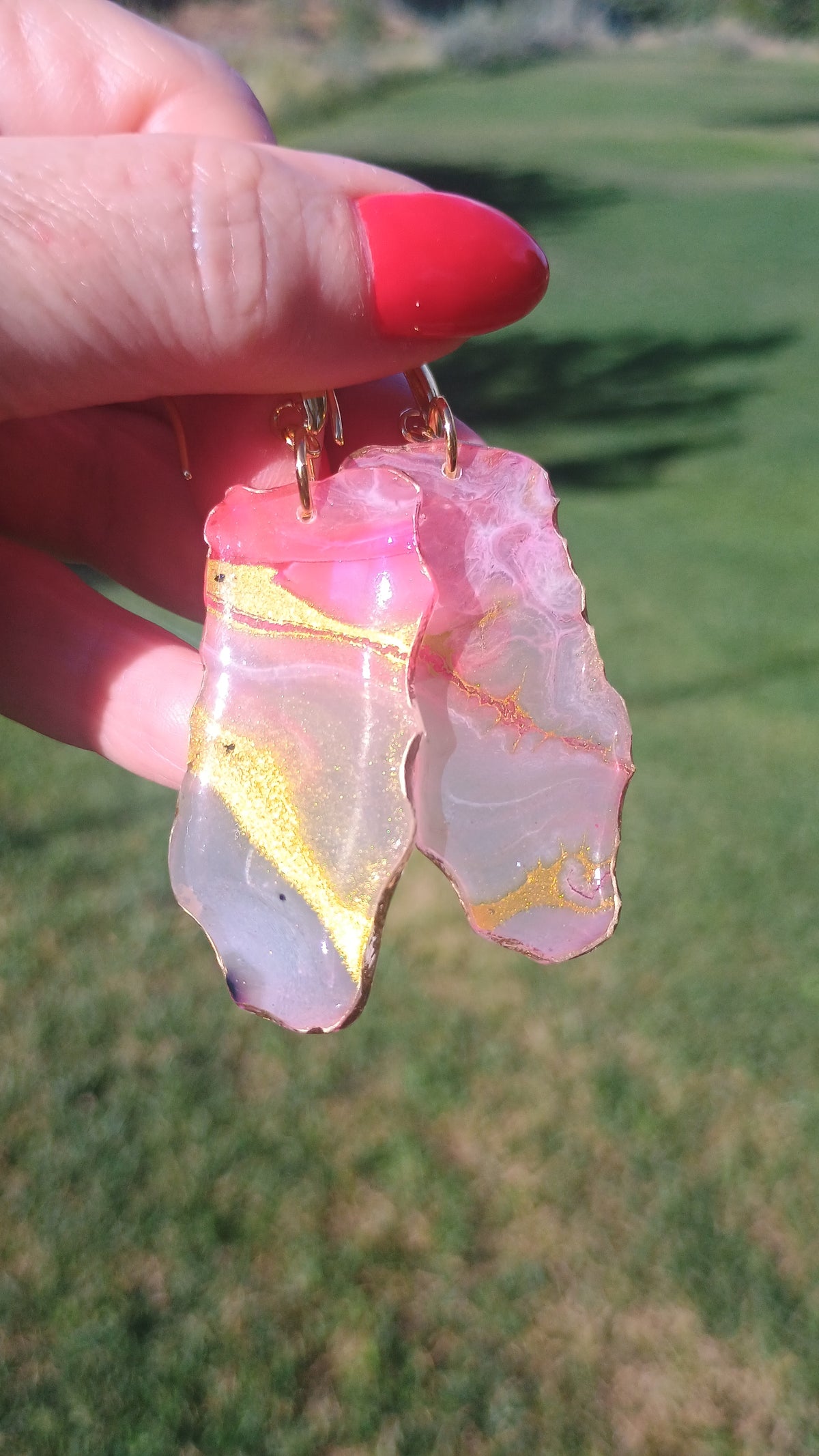 A hand with red nail polish holds a pair of dangling earrings made from pink and white agate looking slices that are actually resin with gold veins, set against a blurred green grass background.