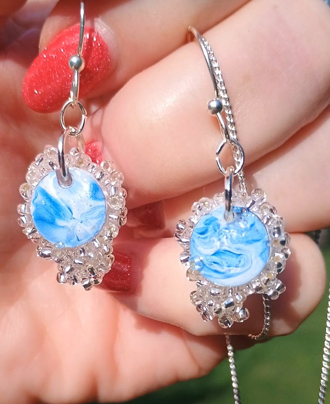 A close-up of a hand holding a pair of dangling earrings and a matching necklace. The jewelry features a round light blue marbled stone in a silver-toned halo setting with clear rhinestones, against a blurred green outdoor background.