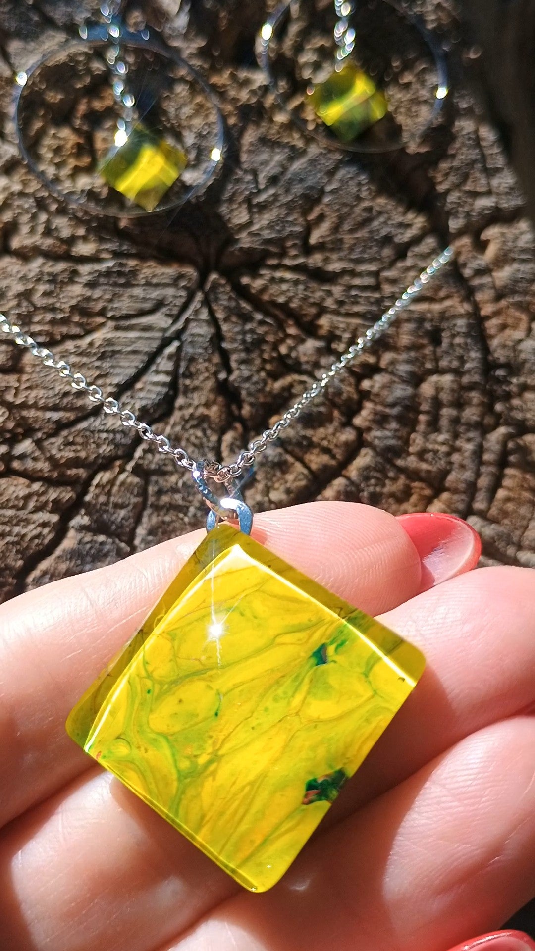 A close-up of a hand holding a square pendant necklace with a bright yellow marbled acrylic painting topped with resin   and a silver chain, with matching dangling earrings blurred in the background on a textured wood surface. A Style by Jess at Gotta Have It Design