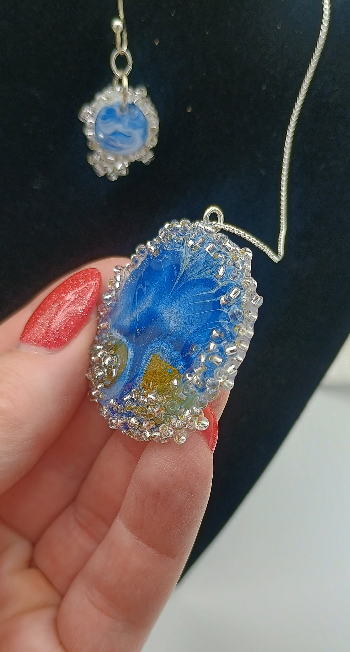 A hand with red nail polish holds an oval pendant necklace featuring a blue and white floral design with a small yellow accent, surrounded by clear rhinestones on a silver-toned setting. A matching necklace is hanging in the background against a black display bust.