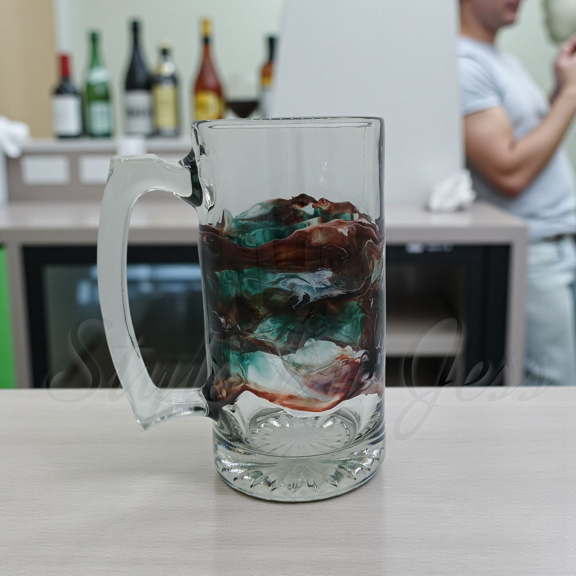 Clear glass mug with handmade art on the outside of the glass on a table, blurred background of a kitchen and bar with individuals socializing. 