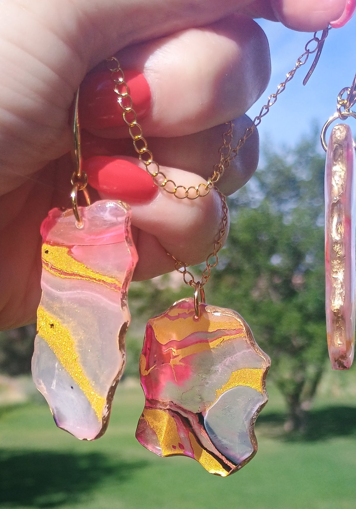A hand with red nail polish holds two gold chain necklaces with irregular, fluid-art glass pendants featuring pink, yellow, and white marbled patterns, set against a blurred outdoor background of green grass and blue sky.