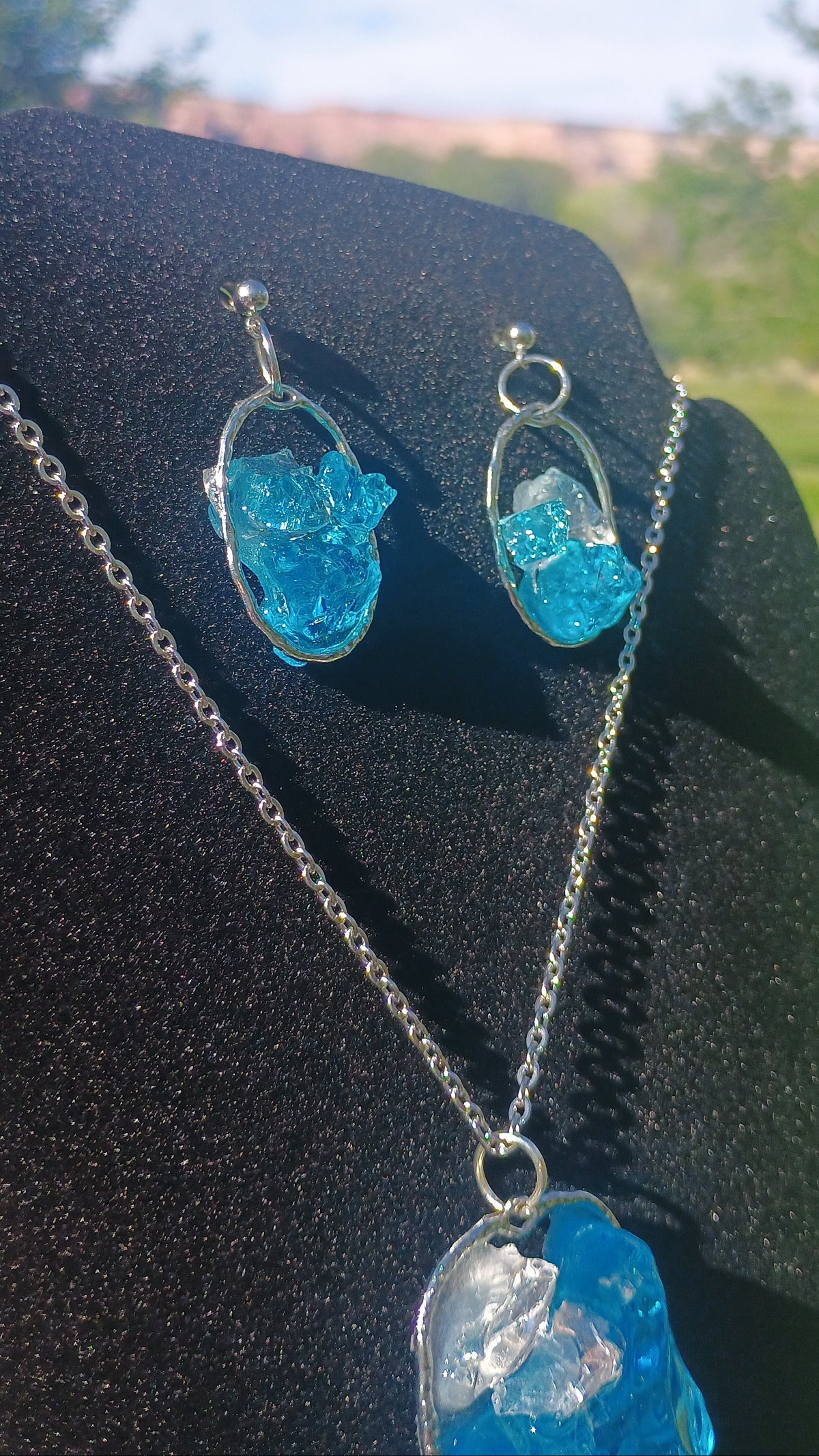 A jewelry set featuring a silver chain necklace and matching stud earrings with irregular, textured bright blue gemstone pendants, displayed on a black velvet stand against an outdoor blurred background.
