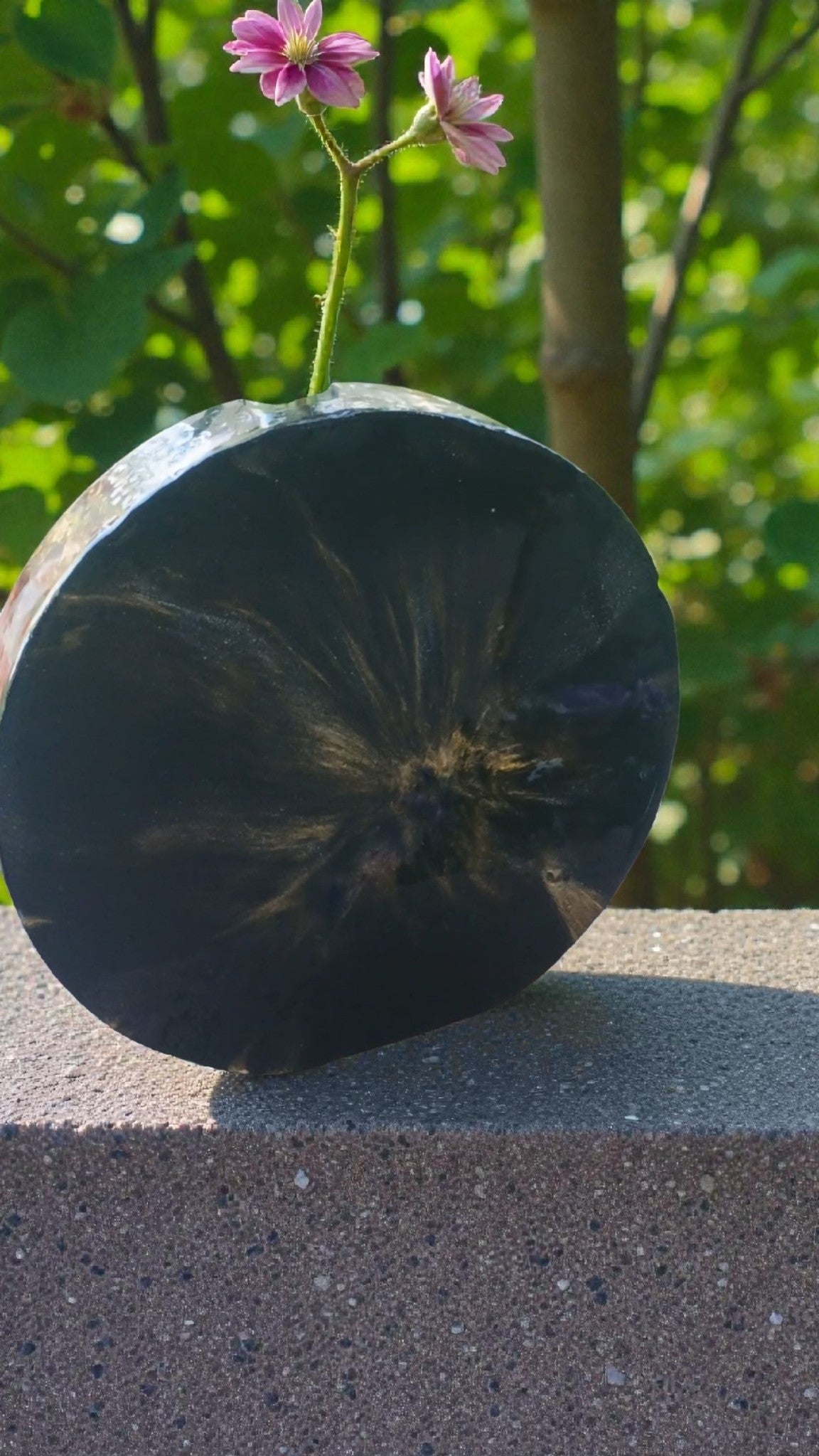 Black resin vase with a natural pattern on a concrete surface with blurred greenery in the background