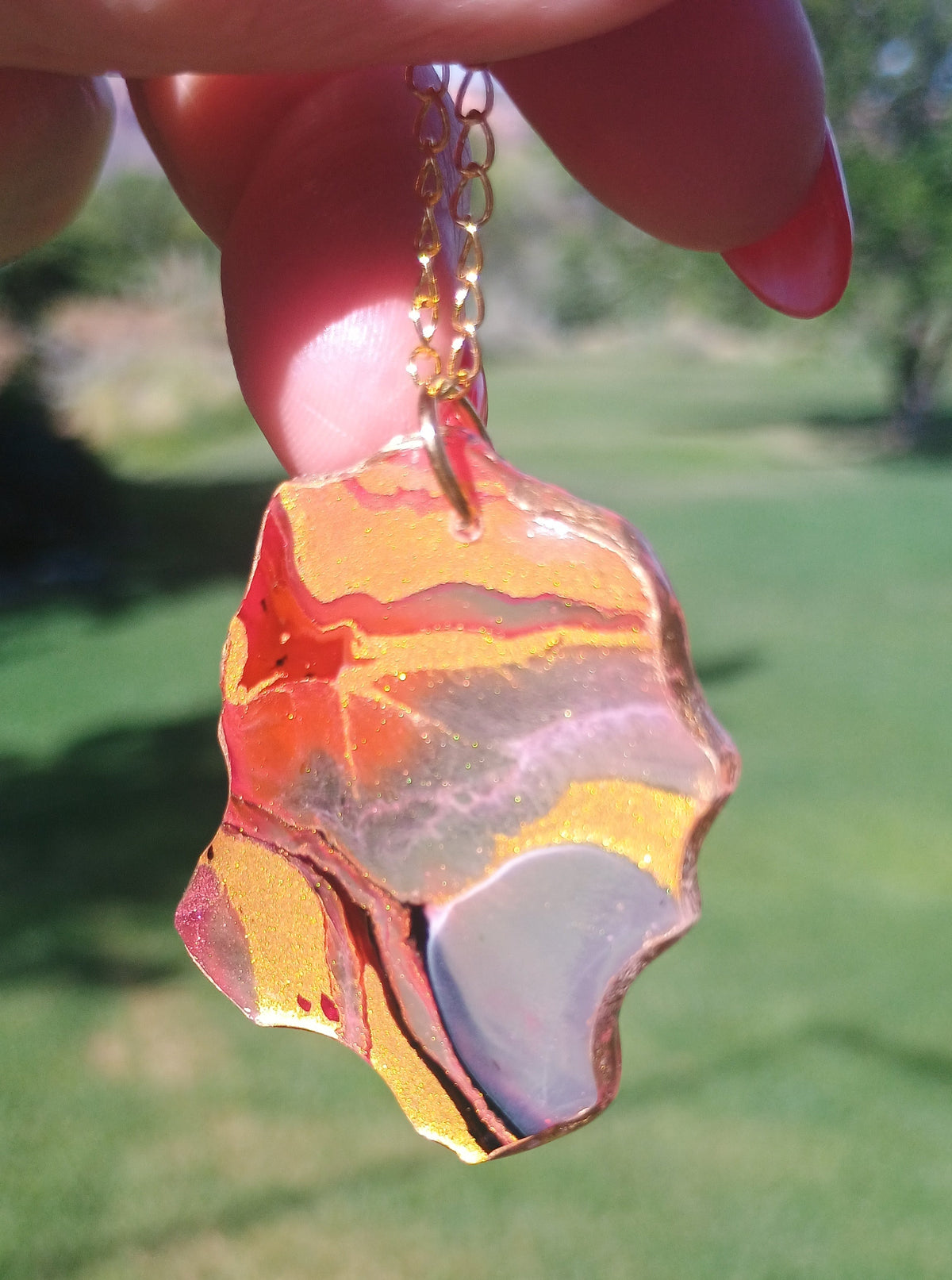 A hand holding a gold chain necklace with a polished, irregularly shaped agate stone looking pendant featuring vibrant pink, gold, red, and gray bands, set against a blurred green grass background.