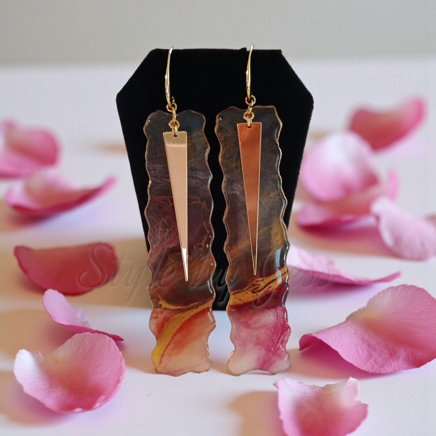 Gold, red and transparent  earrings on a black stand with pink petals around