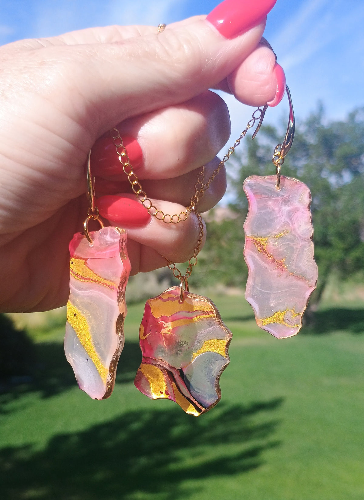 A hand with pink nail polish holds three gold-tone drop earrings featuring irregular, translucent pink agate slice pendants with yellow and white marbling, set against a sunny outdoor background of green grass and blue sky.