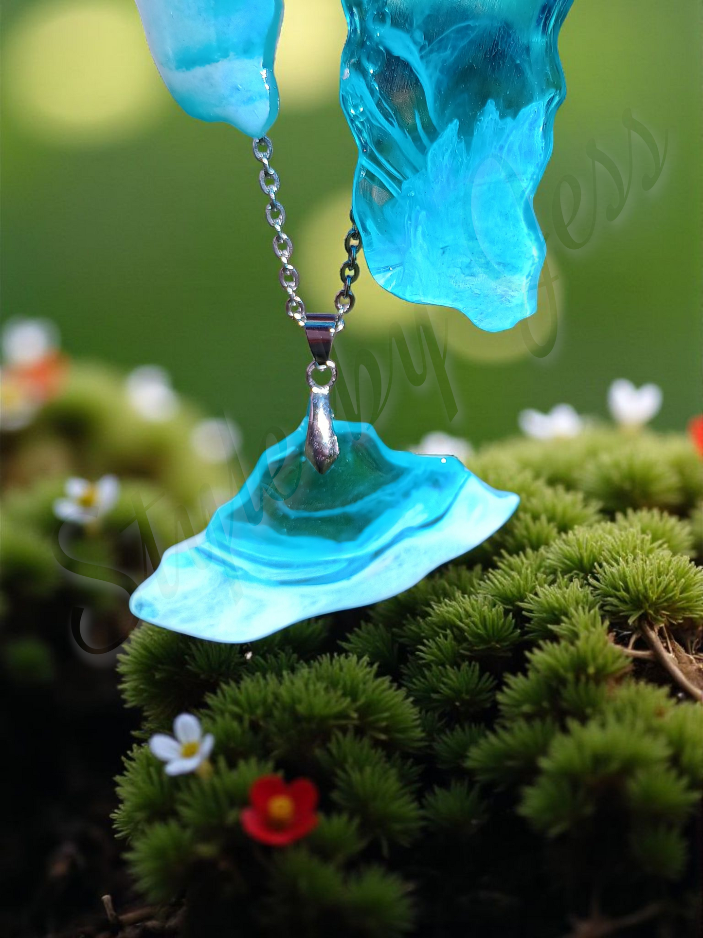 Blue ocean wave pendant hanging on a chain against a blurred green background with flowers.