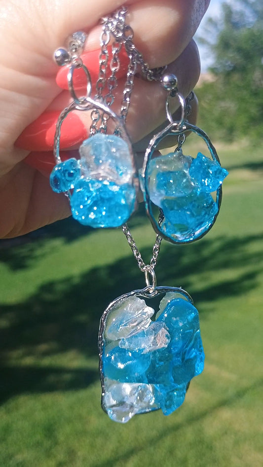 Handmade sea glass jewelry set by Style By Jess — vivid aqua blue and white clear sea glass suspended in transparent clear resin with a floating effect, set in silver-tone oval frames. Includes matching dangle earrings and large pendant necklace with 20-inch silver chain, shown outdoors in natural sunlight against a green background. One-of-a-kind handmade beach jewelry, coastal gift for her. Gotta Have It Design