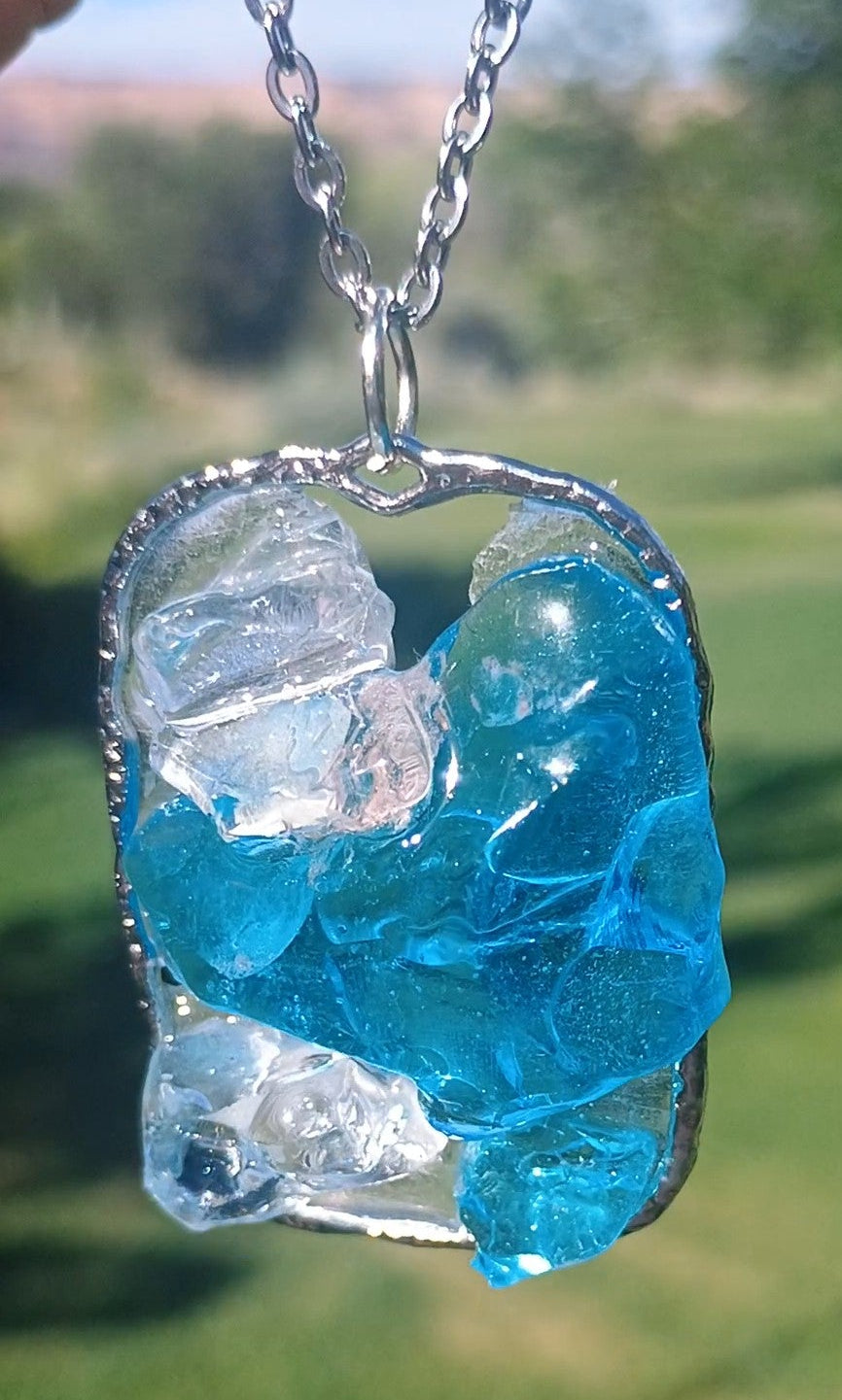 A silver chain necklace with a raw, organic-shaped pendant featuring translucent blue and clear quartz crystals, photographed outdoors against a blurred green grassy background.
