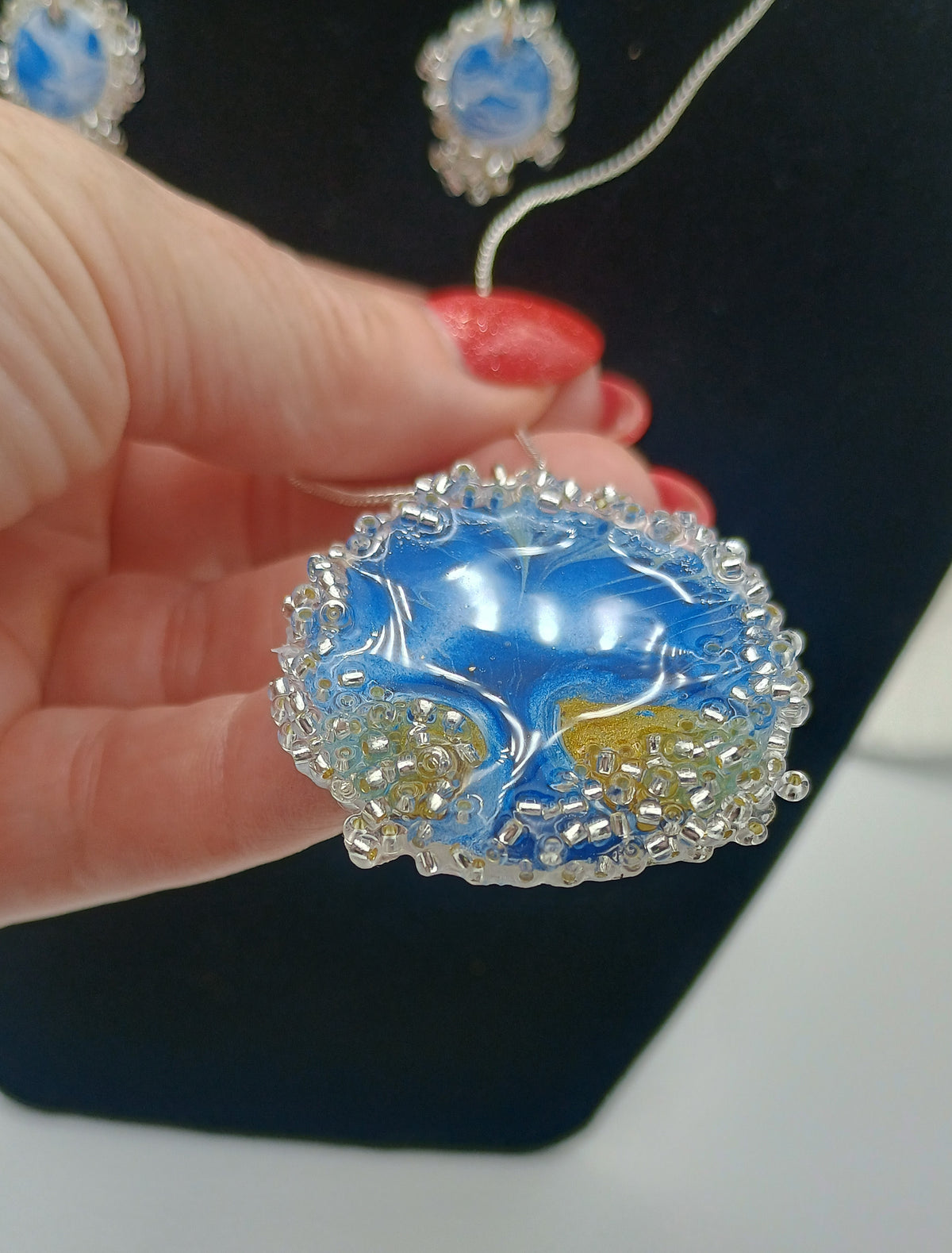 A hand with red painted nails holds a round blue and white tree of life  pendant with a silver textured border. A matching blue and white necklace is draped on a black display bust in the background.