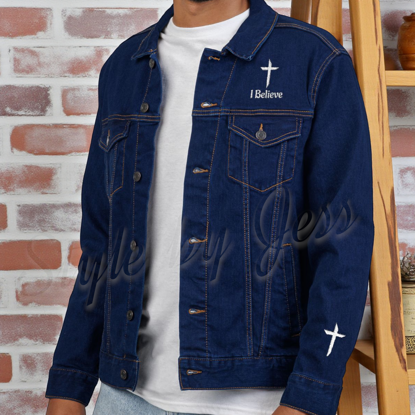 Person wearing a blue denim jacket with 'I Believe' text and cross design against a brick wall.